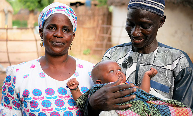 A man and woman stand in front of the camera. The woman looks ahead, the man looks and smiles at their baby in his arms