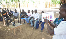 A group of fathers sits in chairs under a tree while one man uses a flip chart of good health behaviors to conduct a training.