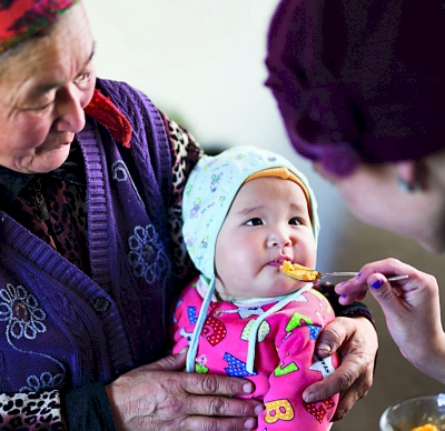 Photo of an infant, held by her grandmother, being fed with a spoon by her mother 
