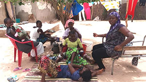 A group of women and children sit in chairs and on the ground outside. They are participating in a household interview.