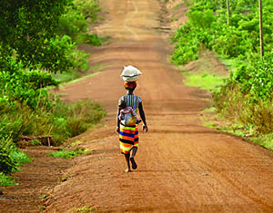 Photo of a woman walking down a long dirt road carrying a package on her head and her child on her back.