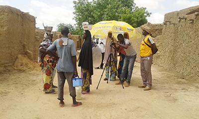 Photo of a video production crew filming two women with children carrying baskets on their head.
