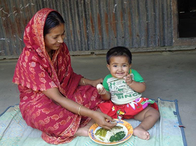 Photo of a mother feeding a her child from a plate set in front of them on a blanket. 