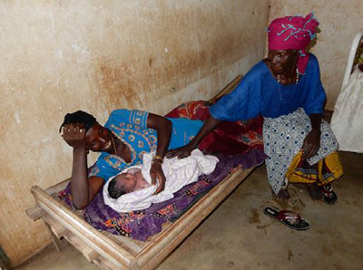 A mother lying on a cot with her child as an older woman/relative sits with them.