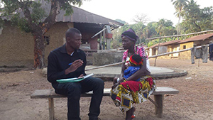  a woman holding her child sits on a bench talking with a man holding a pen and paper.