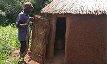 A man stands outside a round latrine structure with a thatched roof.