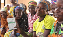 Photo of a woman, Sanatu Fuseini, displaying her VSLA passbook at a meeting with a large group of other women.