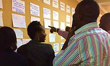 Photograph of several women and men looking at notes posted on a large bulletin board and having a discussion. Caption: "CHAIN partners reflect on joint activities they developed during a November 2016 workshop."