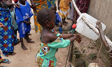 Photo of a child washing her hands with a tippy tap as several other children look on. Caption: "Children in the village of Touba Mourid have fun practicing proper hygiene by washing their hands at a special handwashing station or tippy tap."