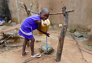 A young child grins at the camera as he washes his hands at a tippy tap.