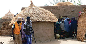 Two men and a woman set up a shot in front of a hut with a video camera. A group of villagers watches from between the hut and a straw wall.