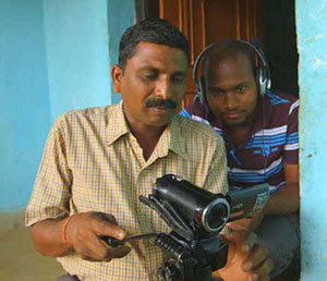 Two men sit together, one behind the other. The man in front holds a video camera and looks at it intensely. The man behind looks wears headphones and has a laptop in his lap.