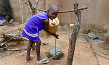 A young child grins at the camera as she washes her hands at a tippy tap.