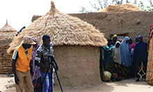 Two men and a woman set up a shot in front of a hut with a video camera. A group of villagers watches from between the hut and a straw wall.
