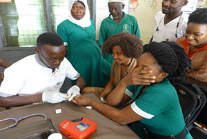 A health worker calmly uses a Hemocue device on a patient. The patient covers her face with her hand and has a pained look on what we can see of her face. Two friends of hers look on with similar uncomfortable looks.