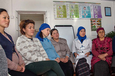A group of six Kyrgyz women sits in a line of chairs.