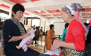  SPRING/Kyrgyz Republic. Two women sitting and talking in a large room with others doing the same. One woman holds cards about nutrition and is counselling the other women who is holding a realistic baby doll and practicing the proper way to hold an infant.