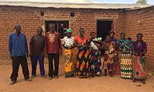 Photo of three men, six women, one girl, and three infants. They are standing outside a brick building, smiling at the camera. Caption: "Proud Mawa beneficiares"