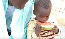 A mother looks on as her child eats a slice of watermelon