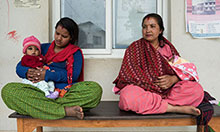 Two Nepali women sit cross-legged on a bench outside what looks like a clinic. One woman holds a roughly 6 month old baby in her lap.