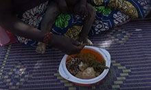 We see the legs of a seated mother and baby with the main focus of the picture on a bowl of diverse fruits and vegetables she is feeding her child.