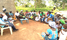 Group photo of a mother-to-mother support group: a large group of women and men sit in plastic chairs in a circle. 