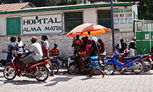 outside of a hospital. people walk by, ride bikes. there is a group of people under an umbrella.