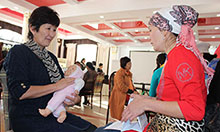 Photo credit: SPRING/Kyrgyz Republic. Two women sitting and talking in a large room with others doing the same. One woman holds cards about nutrition and is counselling the other women who is holding a realistic baby doll and practicing the proper way to hold an infant.