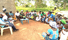 Photo of about a dozen women with their children and several facilitators sitting outside in chairs for a mother-to-mother support group meeting in Ghana