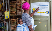 Photo of a mother, carrying her child on her back, reading a poster inside a store. Caption: "A mother looks at 'Benefits of fortified foods' poster in Kampala."