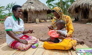 Photo of a woman in a USAID/SPRING shirt sitting on a mat on the ground with a mother holding her child in her arms. The mother is feeding the child with a spoon.